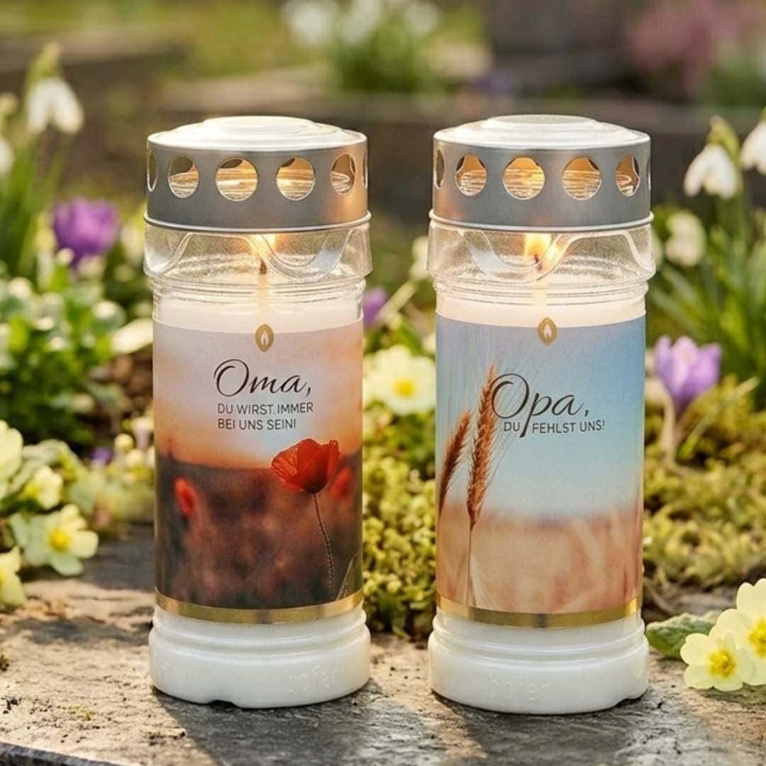 Zwei brennende 5-Tage-Grablichter auf Friedhofsboden, Etiketten „Oma" mit Mohn und „Opa" mit Weizenähren