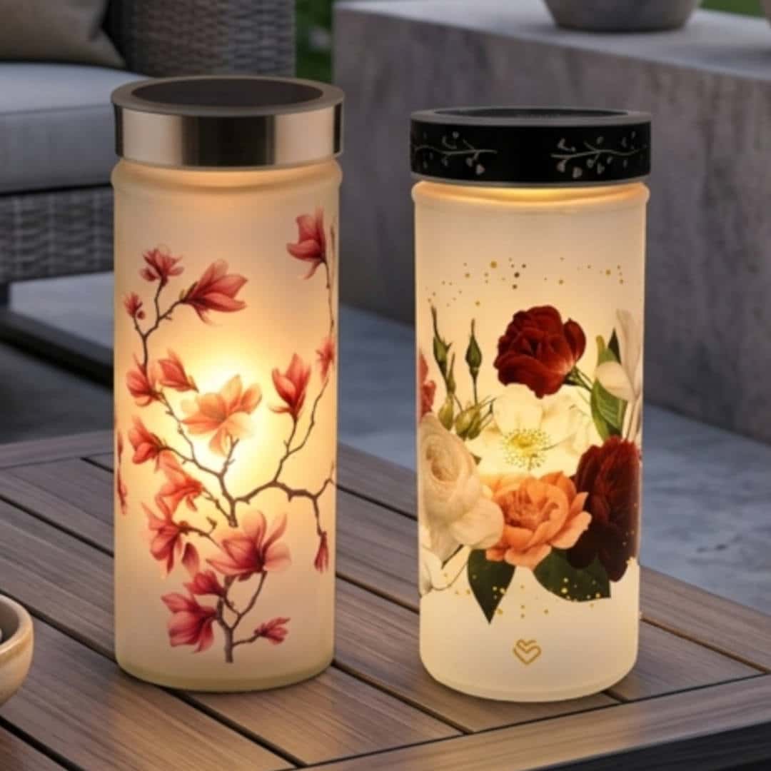 Zwei leuchtende zylindrische Glaslichter auf Holztisch, eines mit Magnolienast, eines mit Rosen- und Blütendekor
