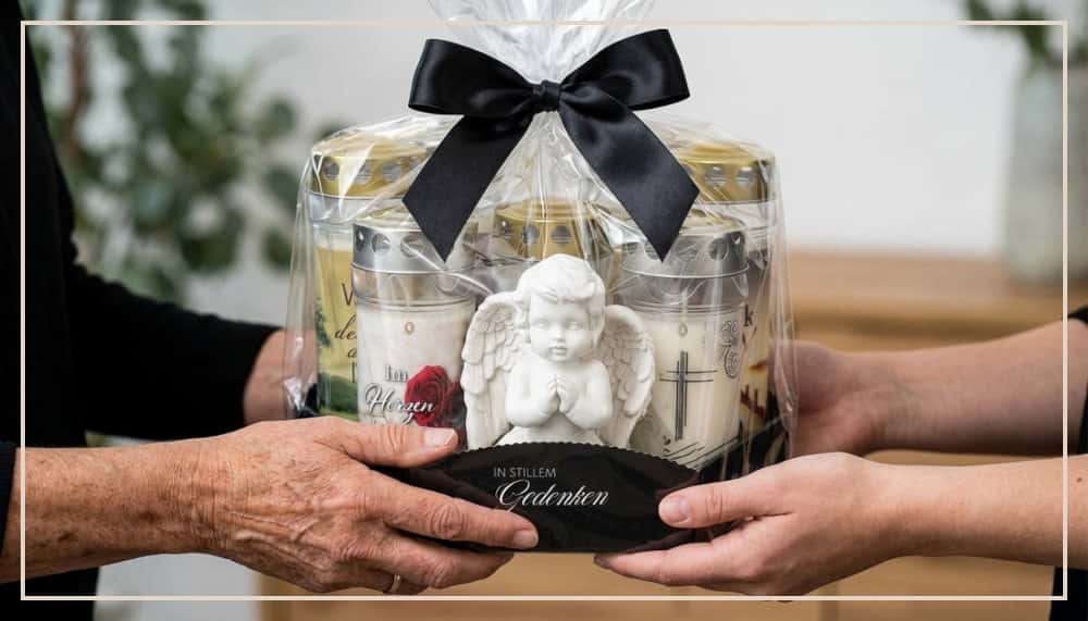 Zwei Personen übergeben ein Trauergeschenk-Set: Mehrere Grablichter mit verschiedenen Motiven, eine weiße Engelfigur und ein schwarzes Tablett mit der Aufschrift „In stillem Gedenken", in Folie eingeschlagen mit schwarzer Satinschleife.